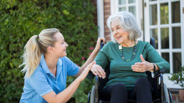 Carer Pushing Senior Woman In Wheelchair Outside Home